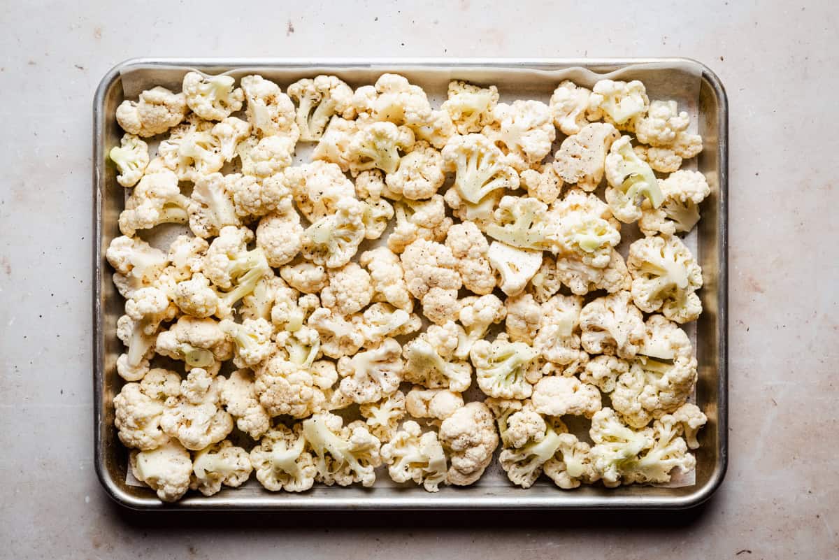 A baking sheet filled with raw cauliflower florets seasoned with spices, ready to be roasted, sits on a light-colored countertop.