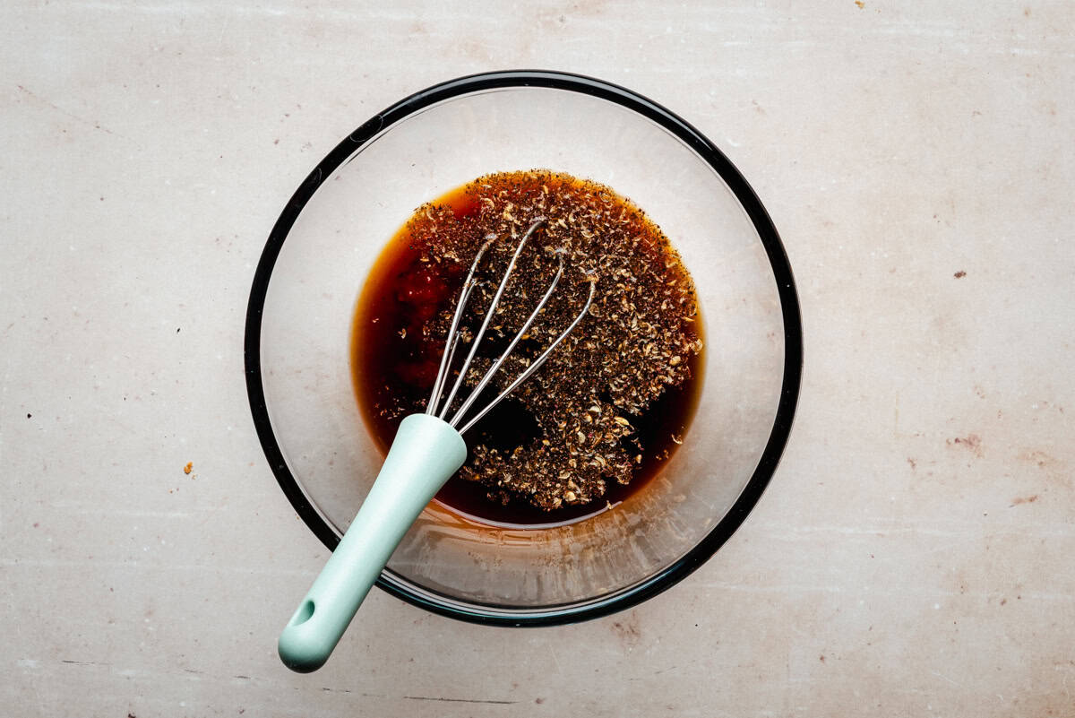 A glass bowl with a light blue whisk mixing a dark liquid with herbs and spices on a light countertop.
