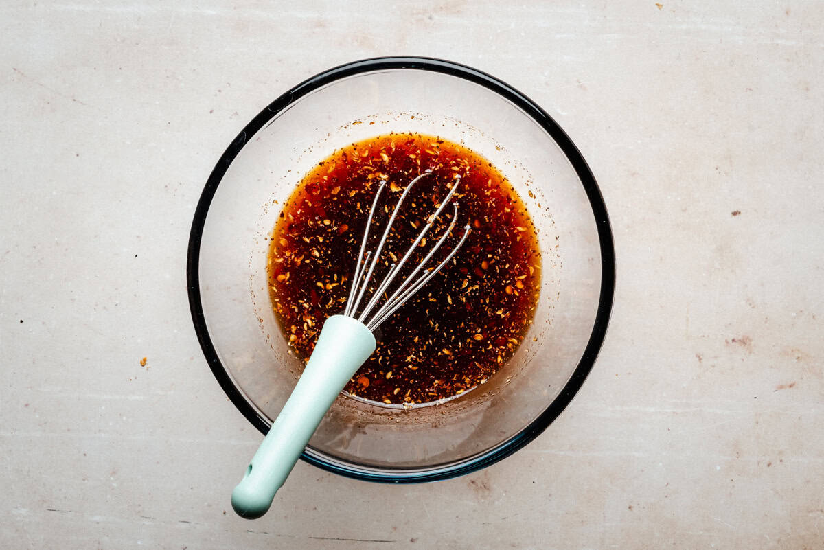 A glass bowl filled with a dark, speckled sauce or marinade, with a whisk resting inside, sits on a light-colored countertop.