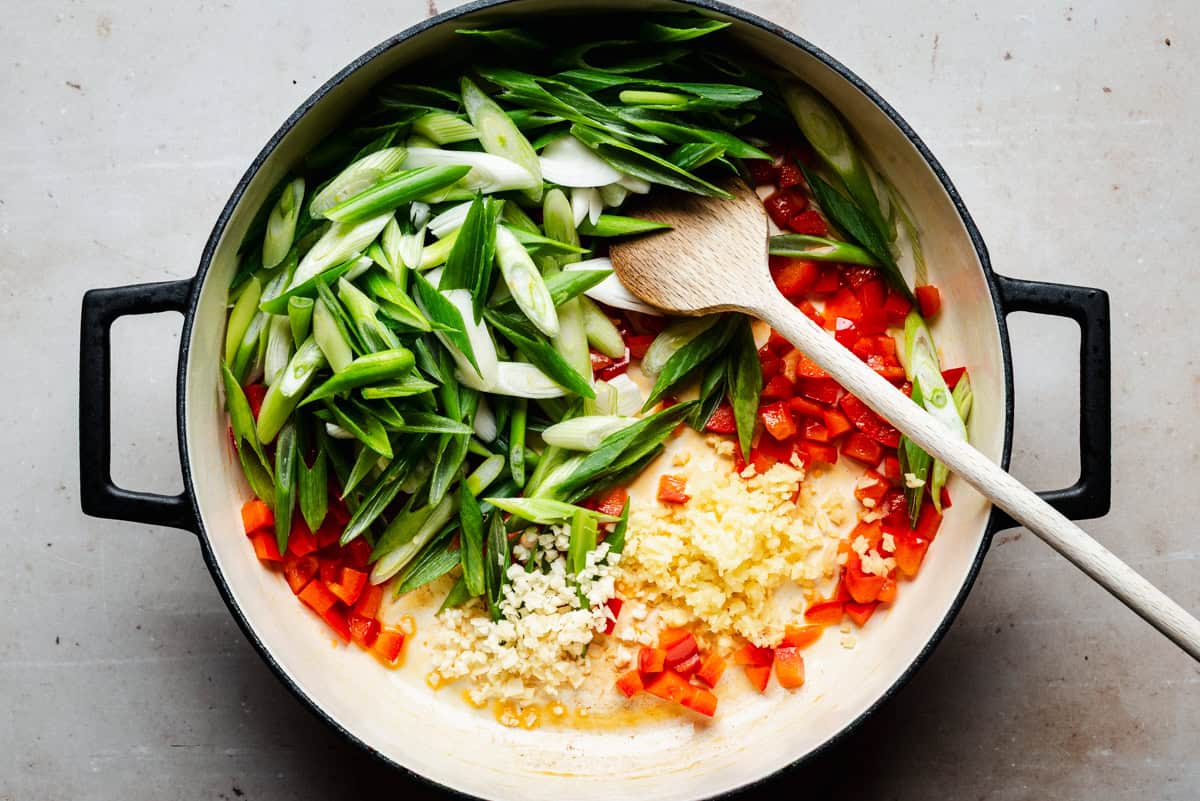 A large pot with sliced green onions, diced red bell peppers, minced garlic, and ginger, with a wooden spoon resting inside, ready for cooking on a light countertop.