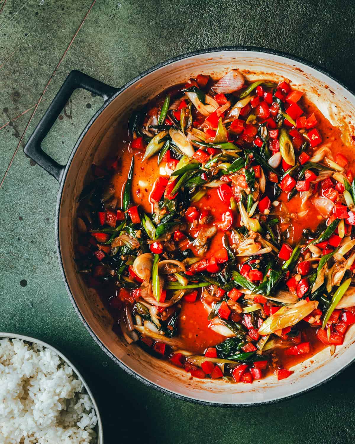 A pot filled with a colorful stir-fry of red bell peppers, green vegetables, and onions in a rich red sauce, next to a bowl of white rice on a green textured surface.