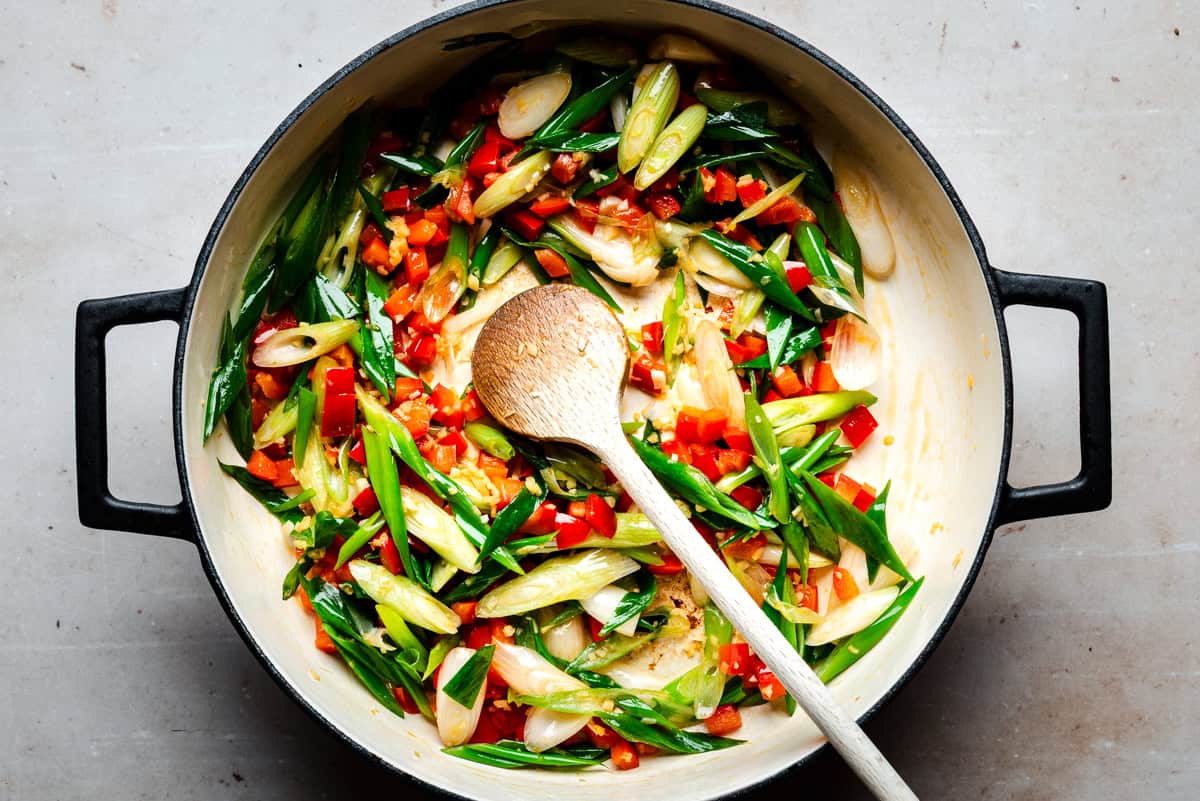 A large pot filled with sliced green onions and red bell peppers being sautéed, with a wooden spoon resting inside. The vegetables are fresh and vibrant against the pot’s light-colored interior.
