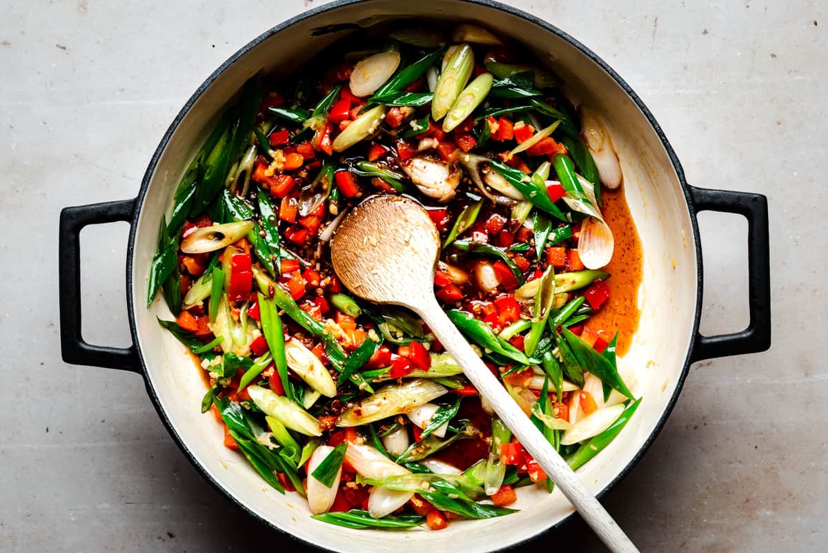 A pot filled kung pao sauce ingredients being stirred with a wooden spoon on a light countertop.
