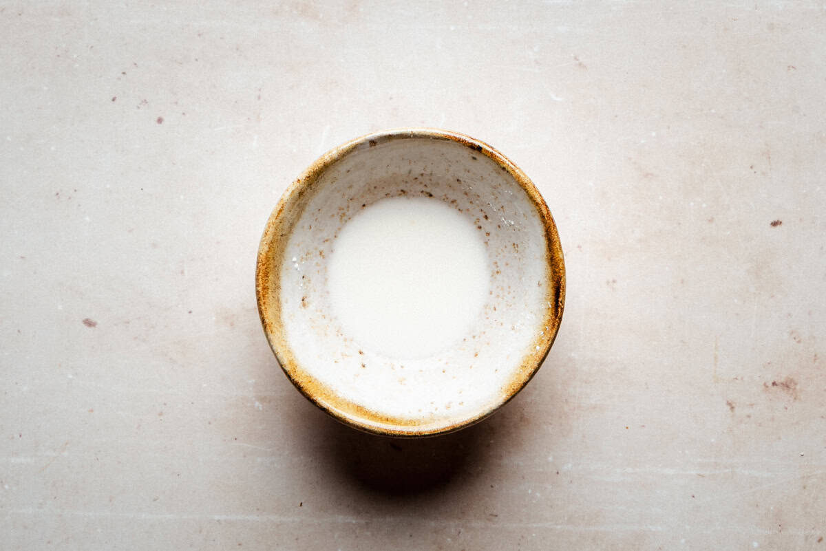A small ceramic bowl with a light brown rim contains a cornstarch slurry, set against a light, textured background.
