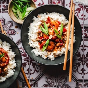 A black bowl filled with white rice topped with stir-fried shrimp, vegetables, peanuts, and green onions, with wooden chopsticks resting on the rim. A bowl of sliced green onions is nearby on a patterned tablecloth.