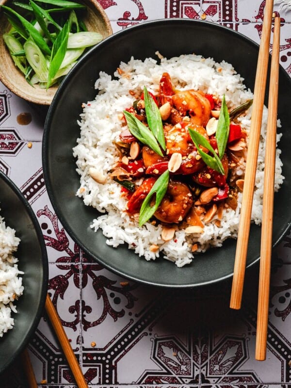 A black bowl filled with white rice topped with stir-fried shrimp, vegetables, peanuts, and green onions, with wooden chopsticks resting on the rim. A bowl of sliced green onions is nearby on a patterned tablecloth.