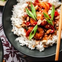 A bowl of white rice topped with Kung Pao shrimp, peanuts, red peppers, green onions, and sauce. A small bowl of sliced green onions is nearby; chopsticks rest on the plate. Text reads "Kung Pao Shrimp.