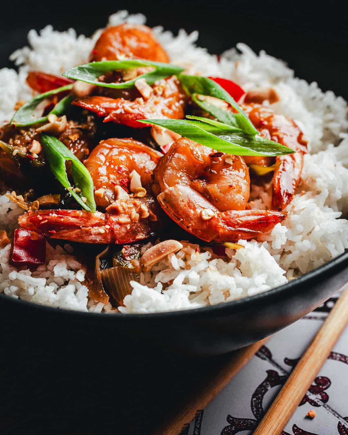 A close-up of a bowl of white rice topped with cooked shrimp in a savory kung pao sauce, garnished with sliced green onions and red chili peppers, with chopsticks resting beside the bowl.