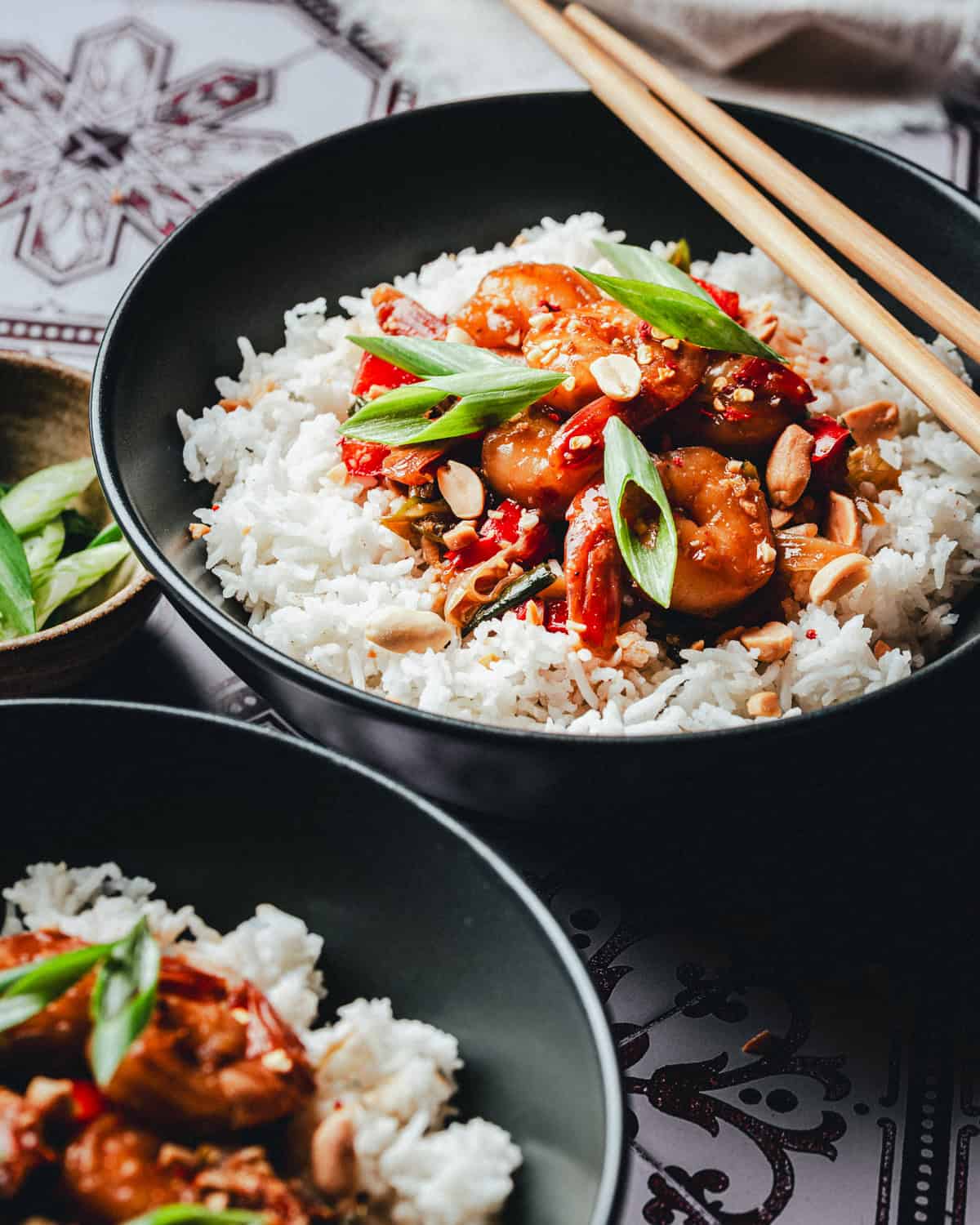 A black bowl filled with white rice topped with saucy kung pao shrimp, sliced green onions, chopped peanuts, and red peppers, with wooden chopsticks resting on the edge. Another bowl with a similar dish is partially visible.