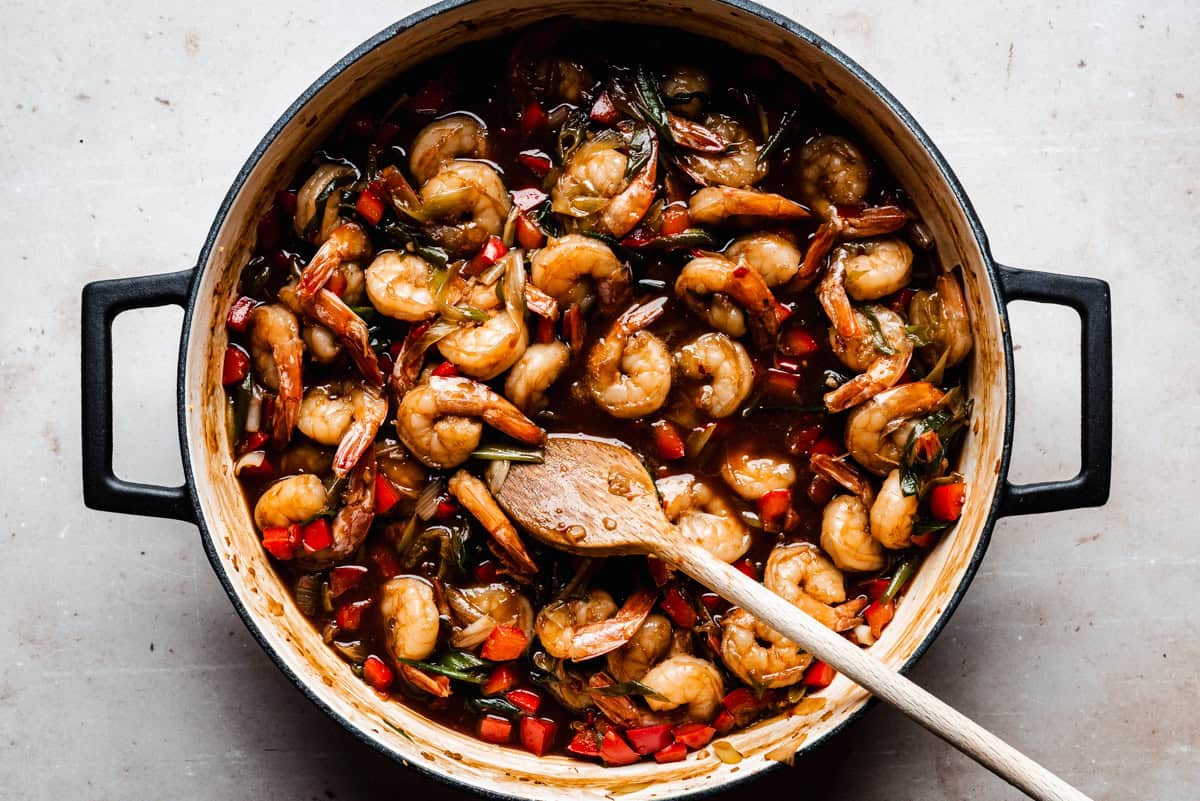 A pot filled with cooked shrimp, red bell peppers, and green onions in a dark, savory sauce, with a wooden spoon resting inside.