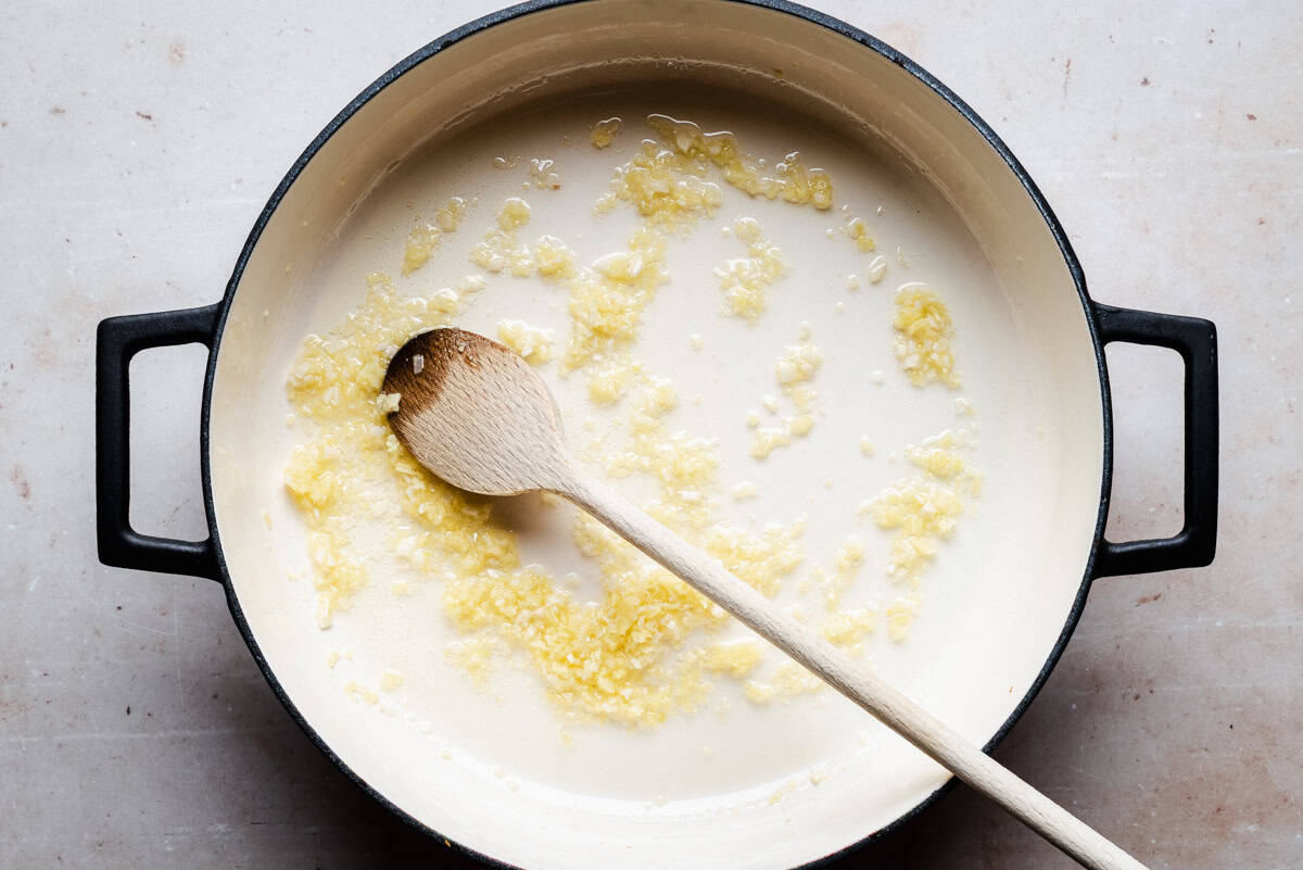 A wooden spoon rests in a pot containing garlic and ginger saut&eacute;ing in oil. The pot sits on a light-colored surface.