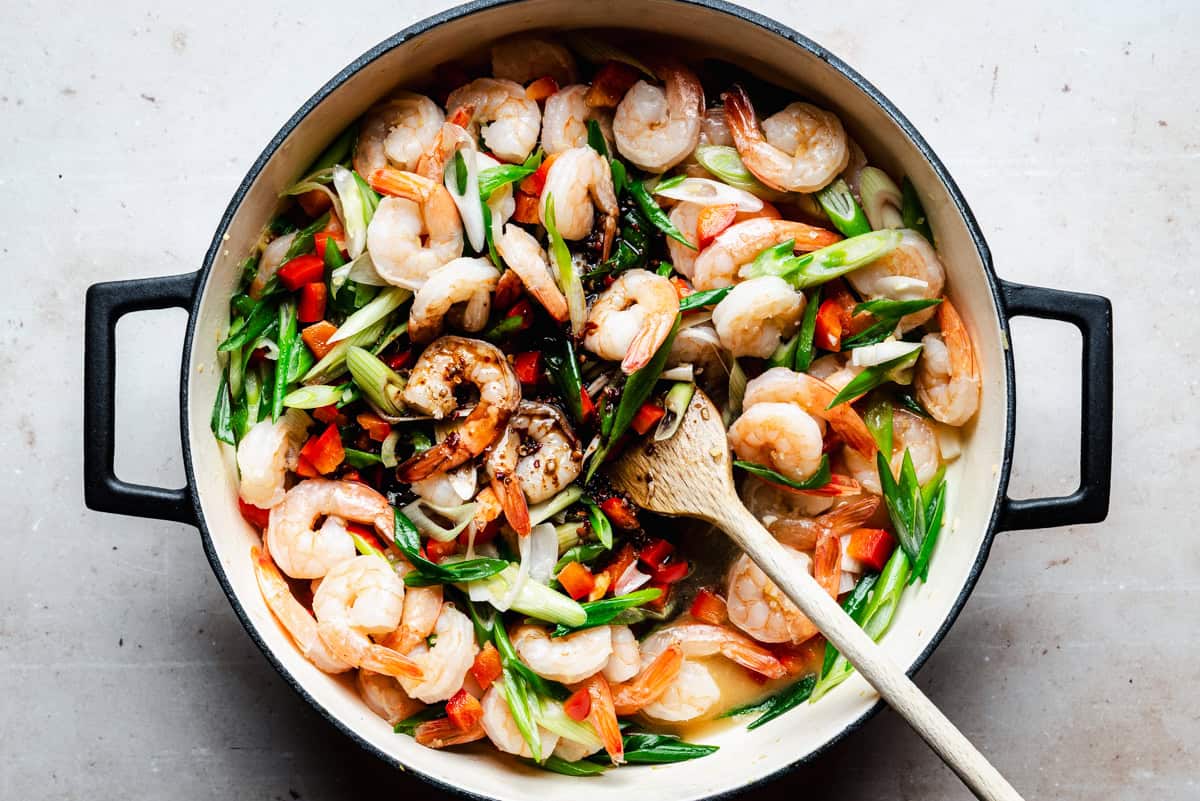 A pot of shrimp stir-fry with sliced green onions, red bell peppers, and a wooden spoon resting inside, viewed from above on a light countertop.