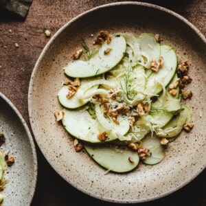 A rustic ceramic plate with a fennel apple salad of thinly sliced green apples, fennel, and chopped walnuts, garnished with fresh dill, set on a brown textured surface.