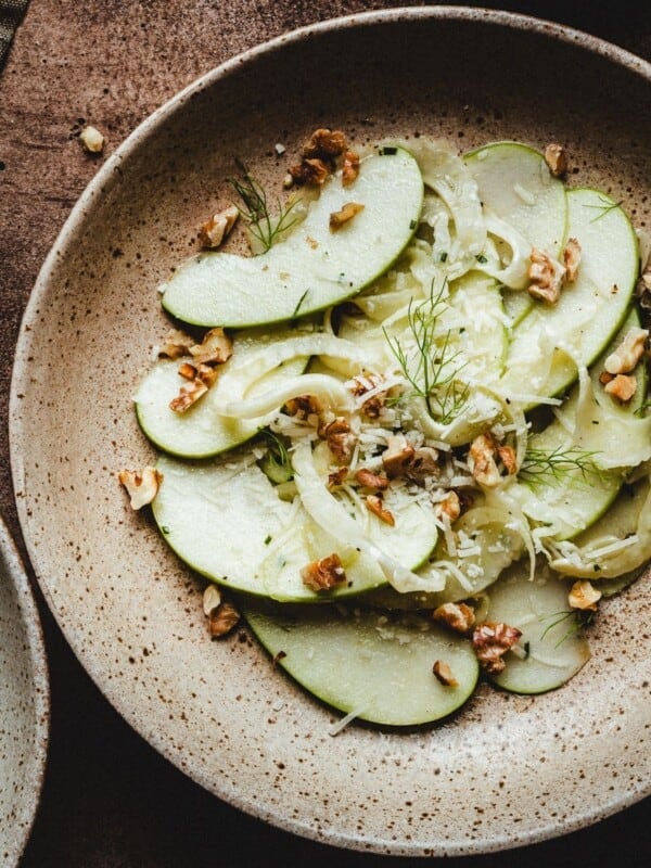 A rustic ceramic plate with a fennel apple salad of thinly sliced green apples, fennel, and chopped walnuts, garnished with fresh dill, set on a brown textured surface.