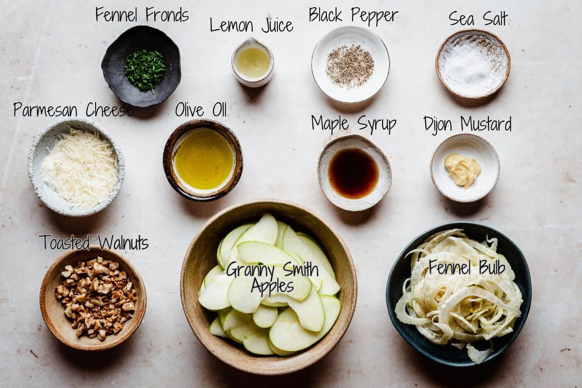 Ingredients for a fennel apple salad arranged on a table, including bowls of fennel fronds, lemon juice, black pepper, sea salt, parmesan cheese, olive oil, maple syrup, Dijon mustard, toasted walnuts, sliced apples, and fennel bulb.