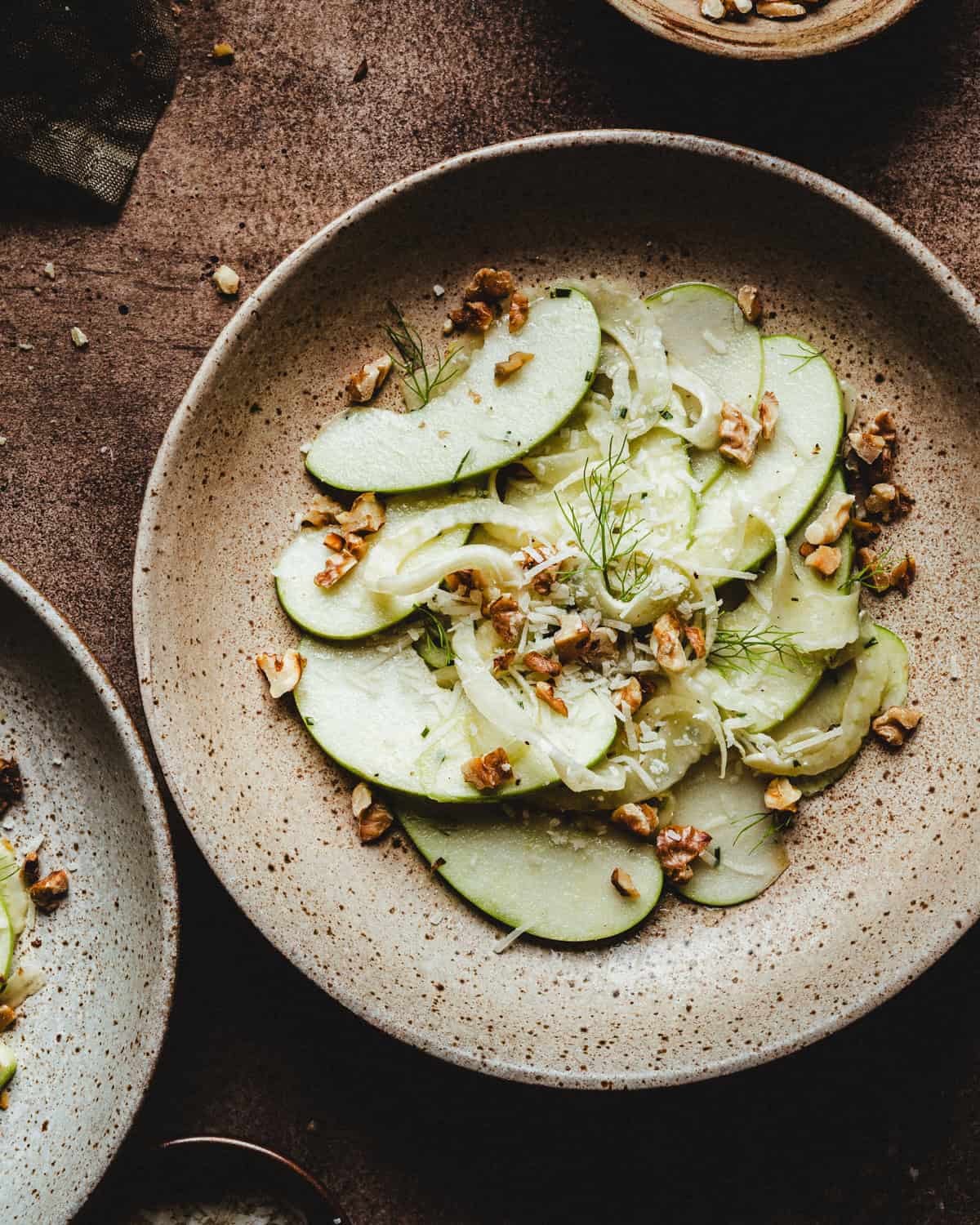 A rustic ceramic plate with a fresh salad made of thinly sliced green apples, fennel, shredded cheese, and chopped walnuts, garnished with herbs. The plate sits on a brown textured surface.
