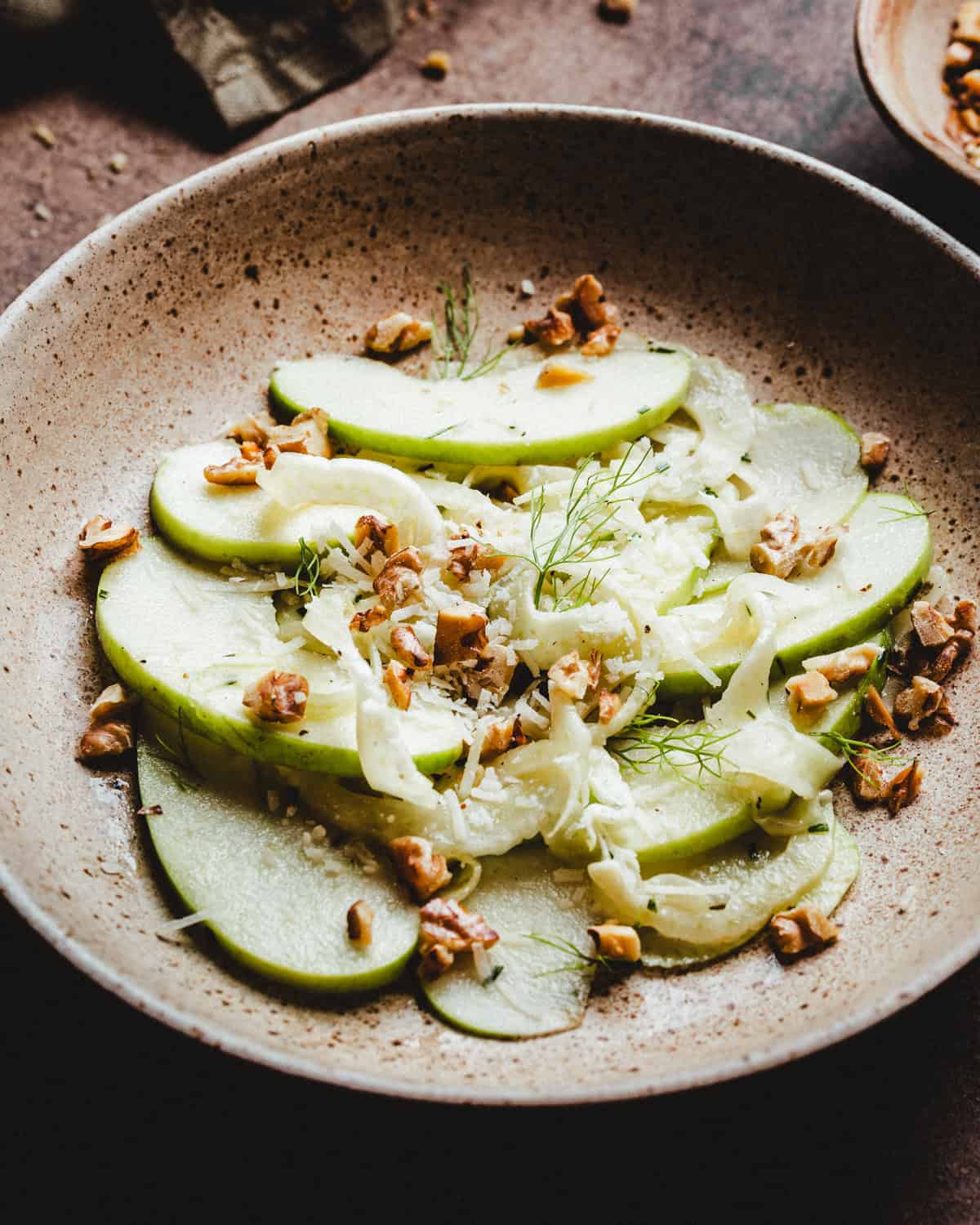 A ceramic bowl filled with thinly sliced green apples, shredded fennel, chopped walnuts on a rustic tabletop.