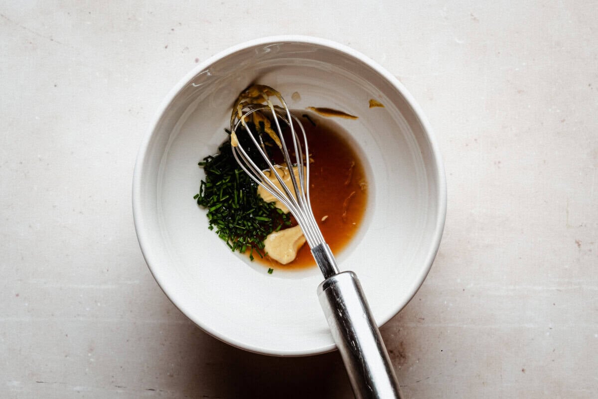 A metal whisk rests in a white bowl containing maple syrup, chopped chives, a dollop of mustard, and a portion of garlic or ginger, ready to be mixed.