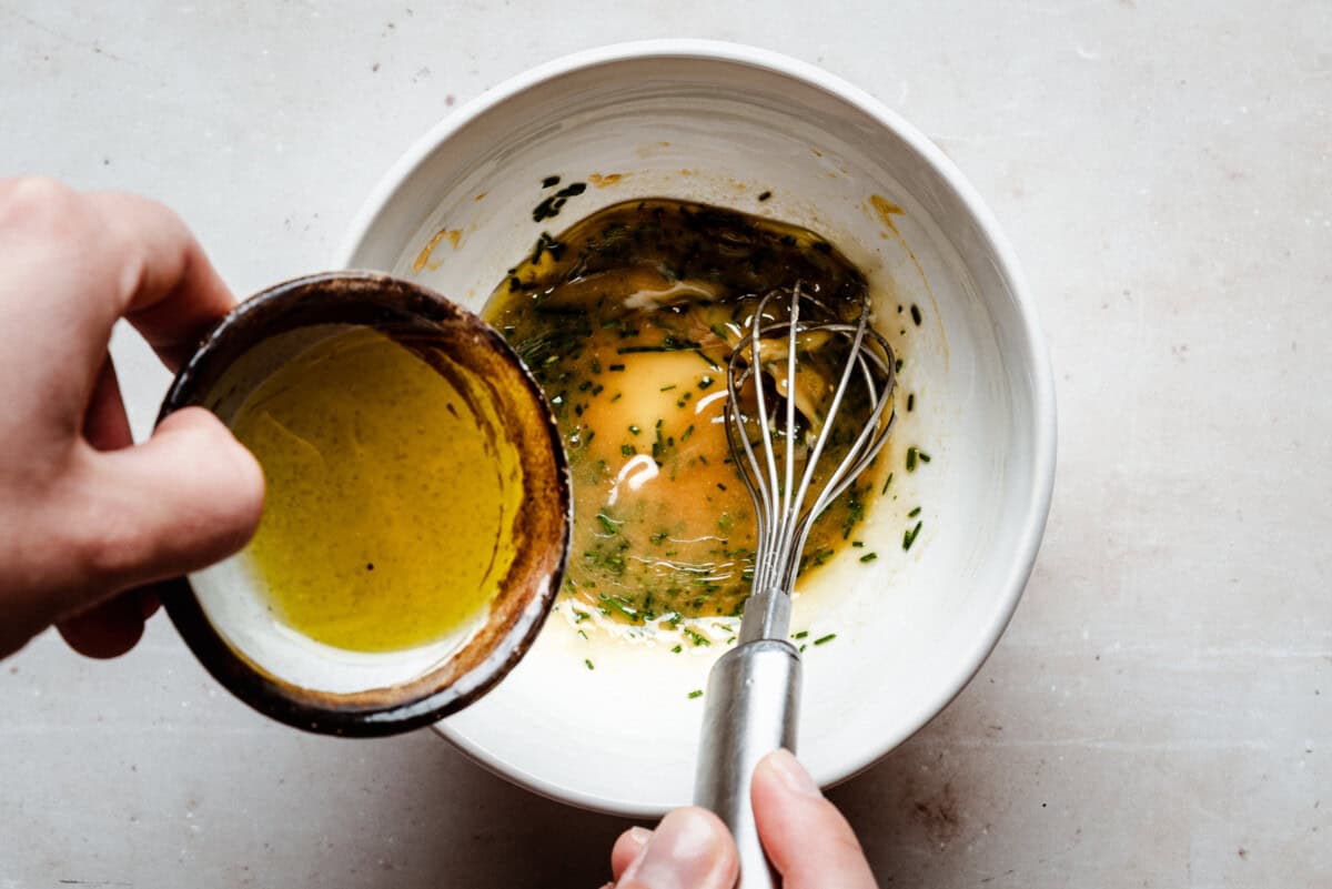 A person pours olive oil from a small cup into a white bowl while mixing the ingredients with a metal whisk.