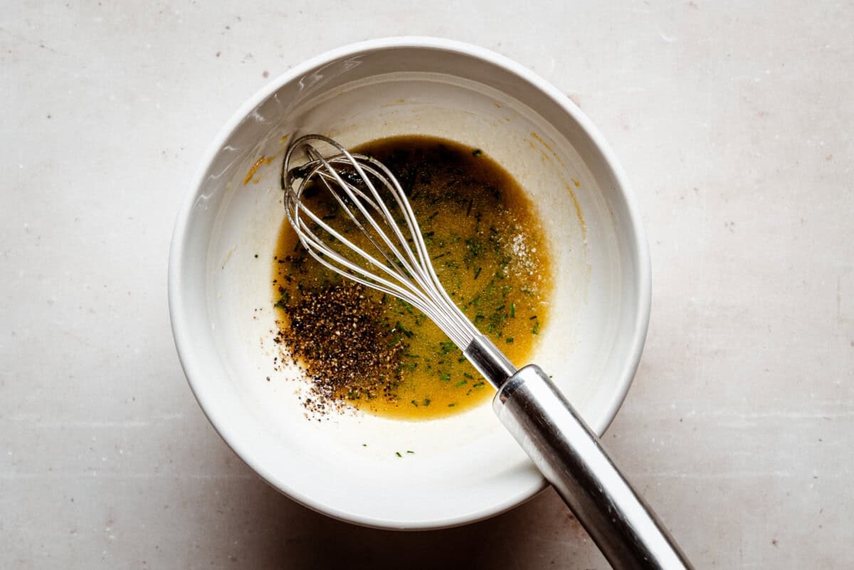 A metal whisk rests in a white bowl containing a mixture of oil, herbs, salt, and ground black pepper on a light countertop.