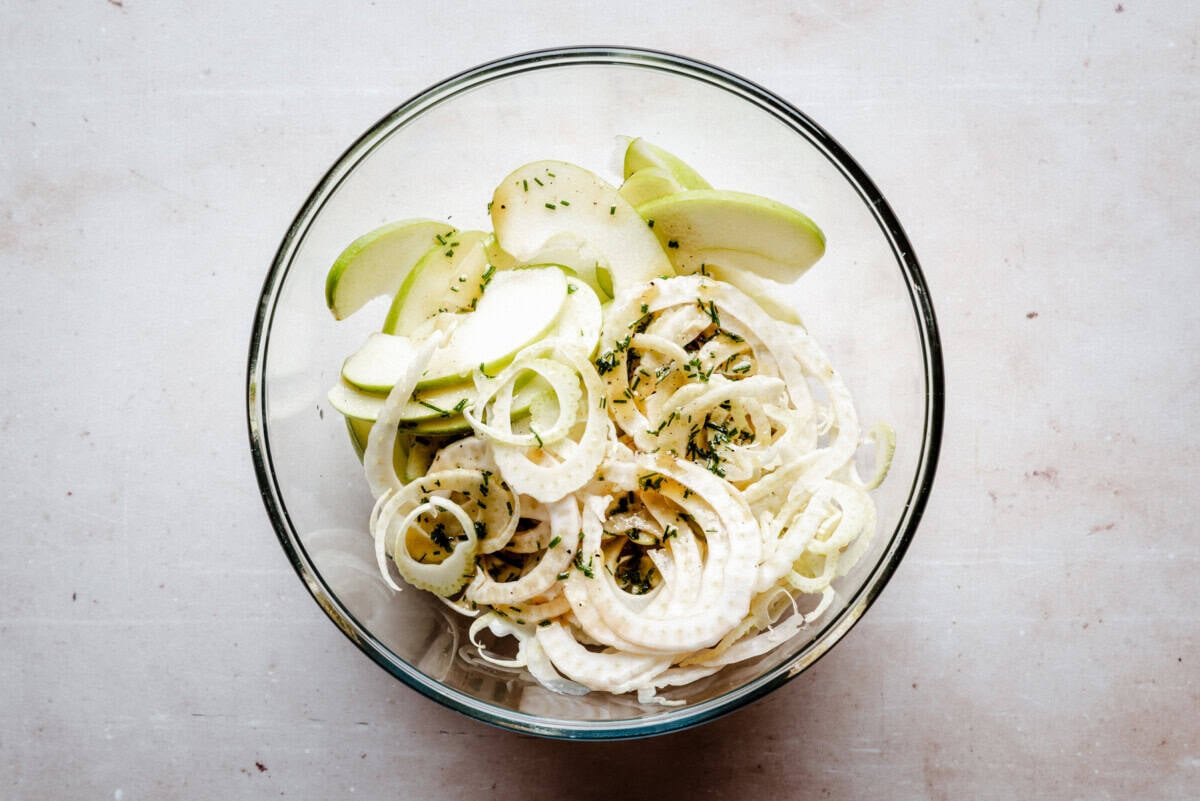 A glass bowl filled with thinly sliced green apples and fennel, sprinkled with herbs, sits on a light-colored surface.