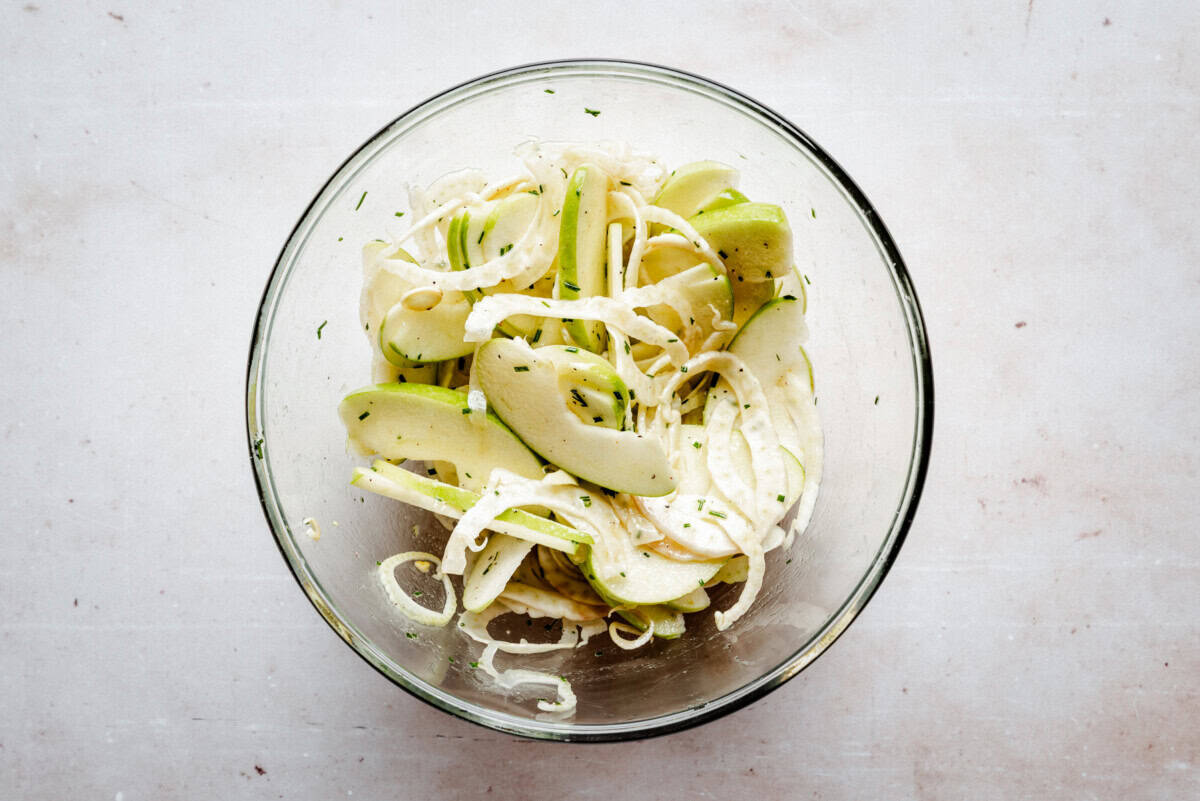 A glass bowl containing a salad of thinly sliced green apples and fennel, mixed with herbs, on a light-colored surface.