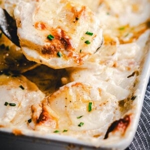 A close-up of creamy, golden-brown scalloped potatoes garnished with chopped chives in a baking dish, with a serving spoon lifting a portion.
