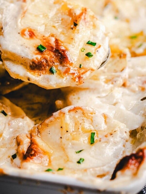 A close-up of creamy, golden-brown scalloped potatoes garnished with chopped chives in a baking dish, with a serving spoon lifting a portion.