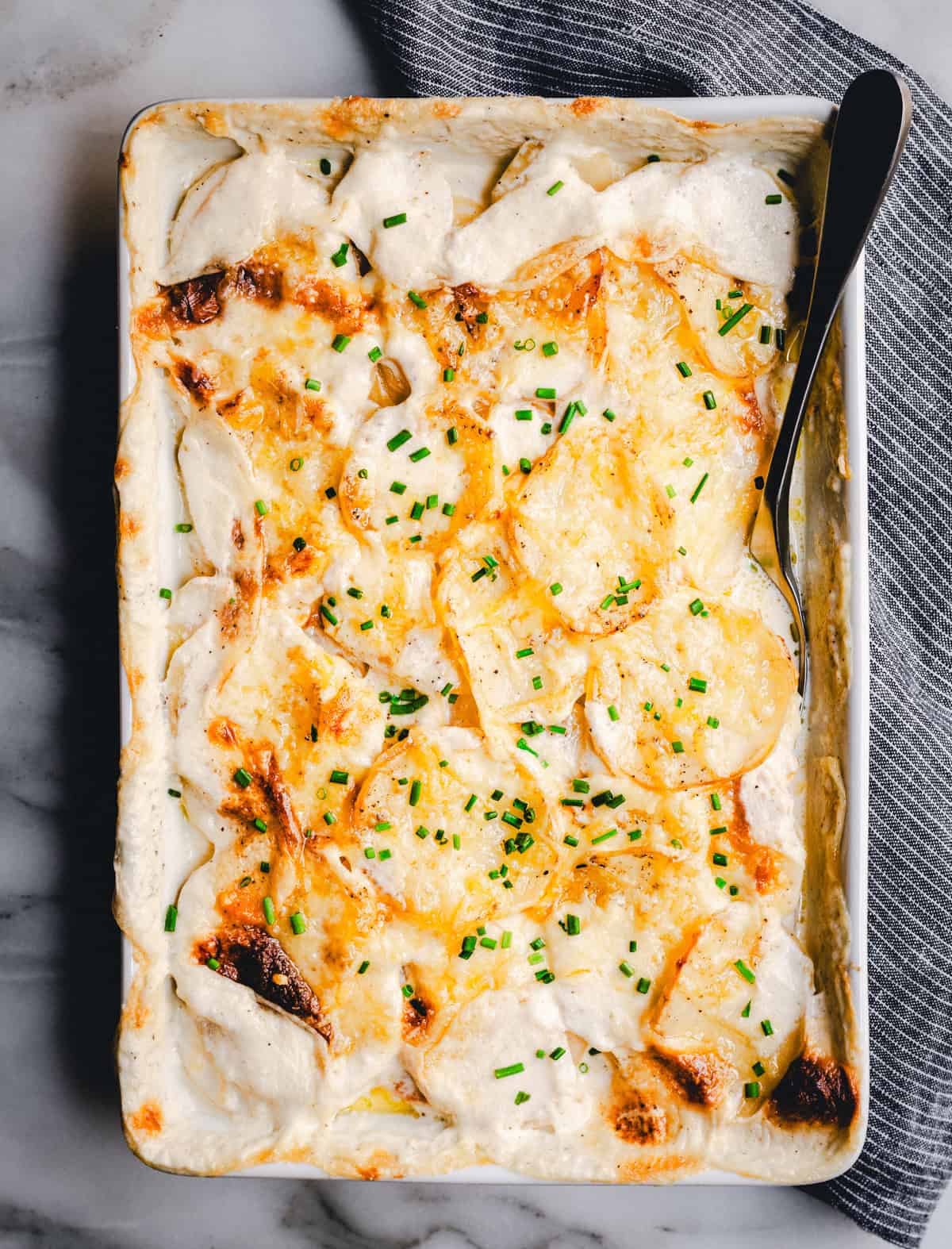 A rectangular baking dish filled with creamy, golden-brown scalloped potatoes topped with chopped chives. A serving spoon rests on the side of the dish, and a gray-striped cloth is nearby on a marble surface.