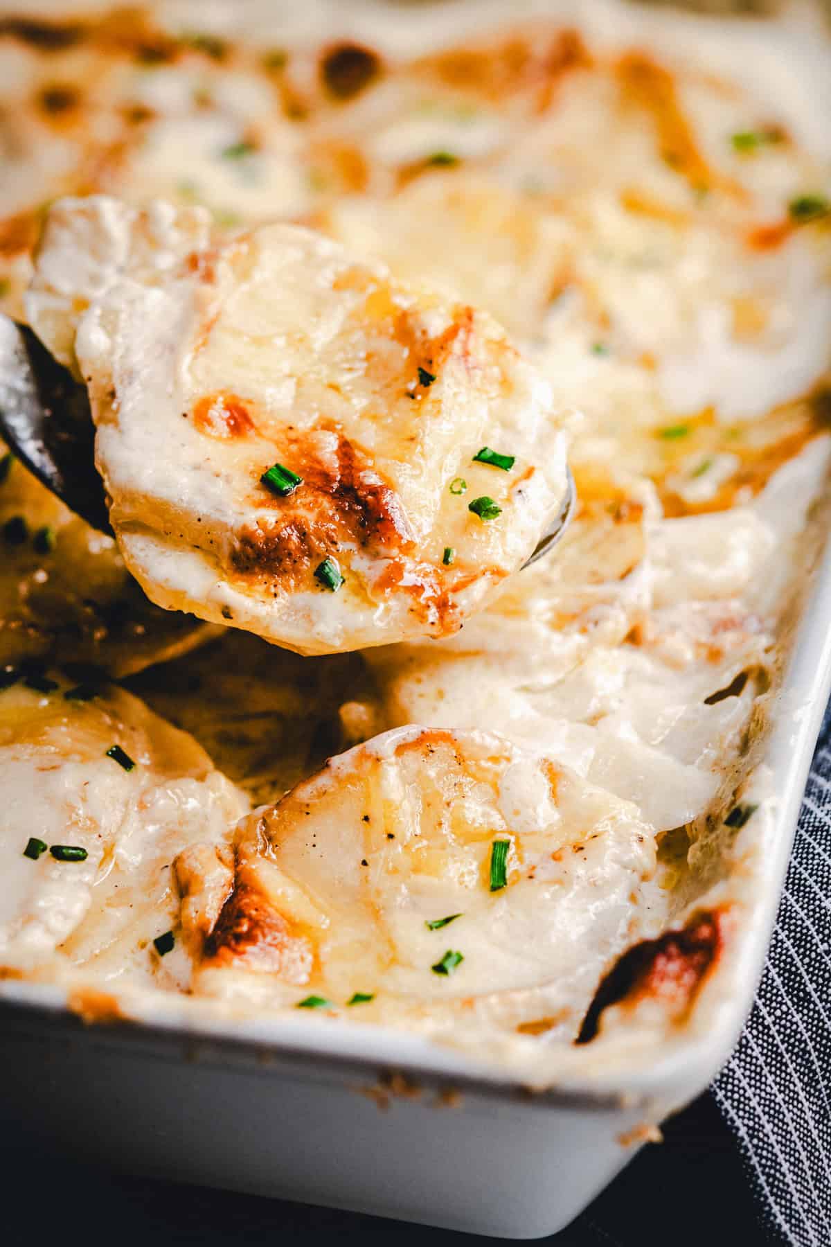 A close-up of creamy, golden-brown gratin dauphinois potatoes being served from a baking dish, garnished with chopped chives.