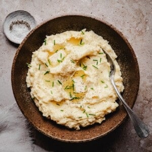 A rustic brown bowl filled with creamy horseradish mashed potatoes, topped with chopped chives, ground black pepper, and a drizzle of melted butter, with a spoon resting inside. A small dish of salt sits nearby on a textured surface.
