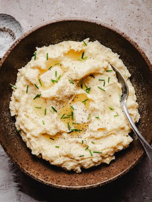 A rustic brown bowl filled with creamy horseradish mashed potatoes, topped with chopped chives, ground black pepper, and a drizzle of melted butter, with a spoon resting inside. A small dish of salt sits nearby on a textured surface.