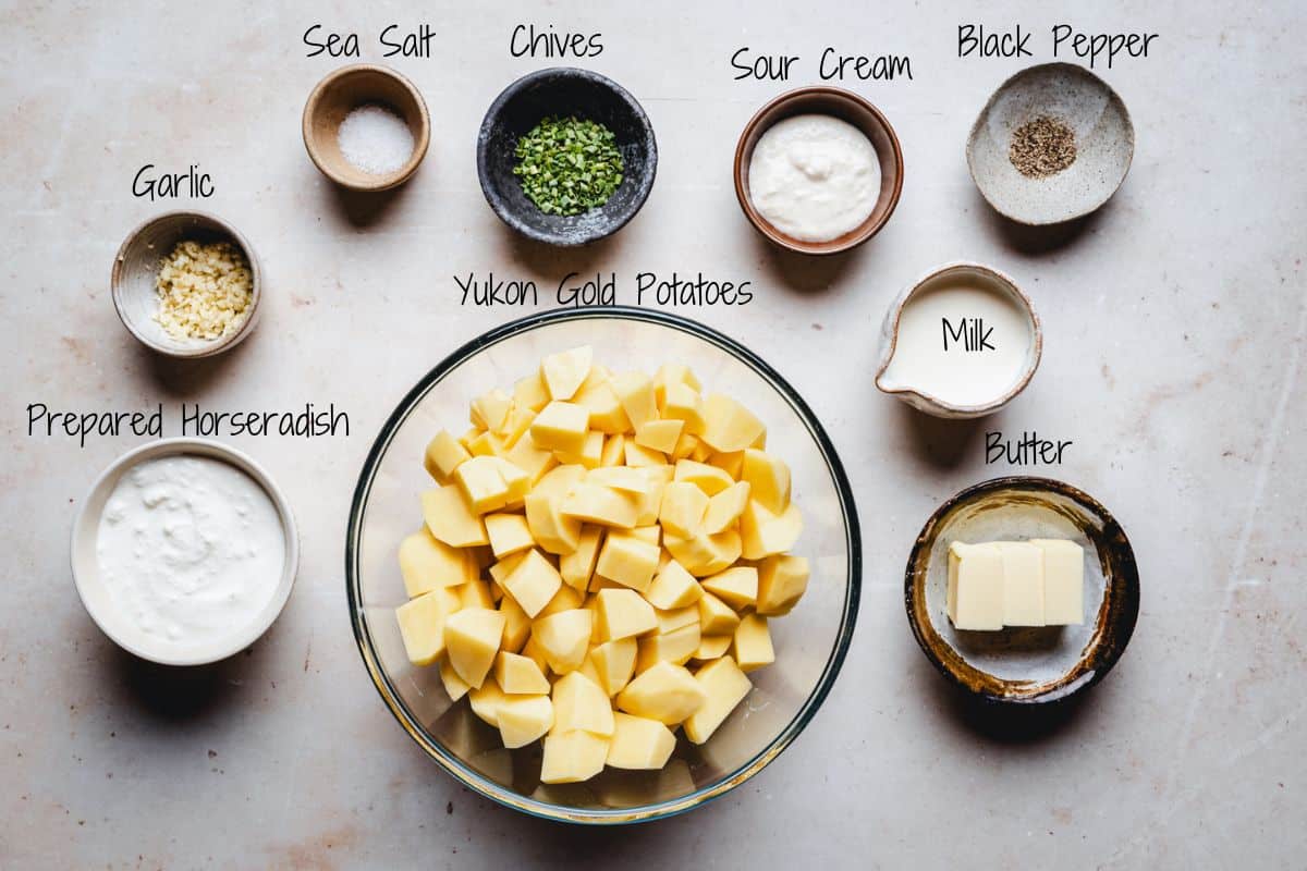 A bowl of cubed Yukon Gold potatoes surrounded by small bowls of sea salt, chives, sour cream, black pepper, minced garlic, prepared horseradish, milk, and butter on a light surface.