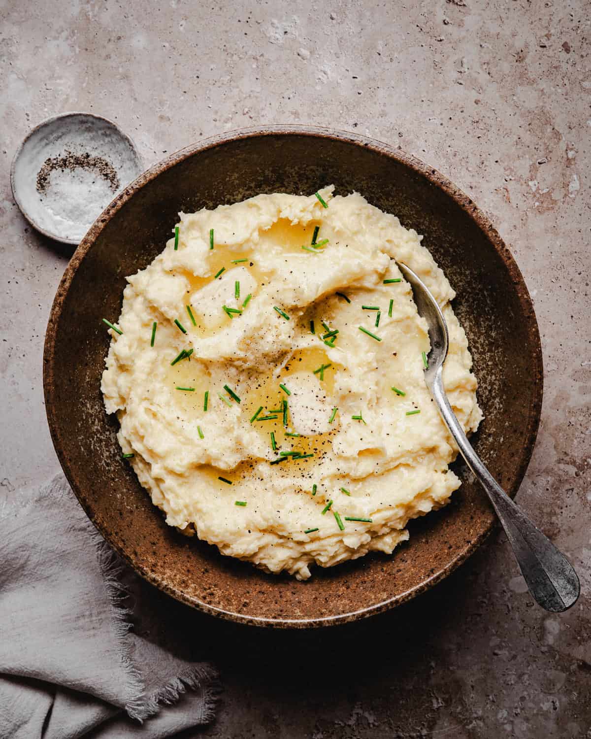 A rustic brown bowl filled with creamy mashed potatoes, topped with butter, chopped chives, and black pepper. A spoon rests in the bowl, with a small dish of salt nearby on a textured surface.