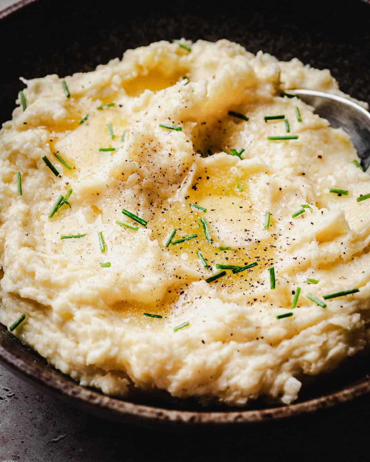 A bowl of creamy mashed potatoes topped with melted butter, chopped chives, and a sprinkle of black pepper, with a spoon resting in the dish.
