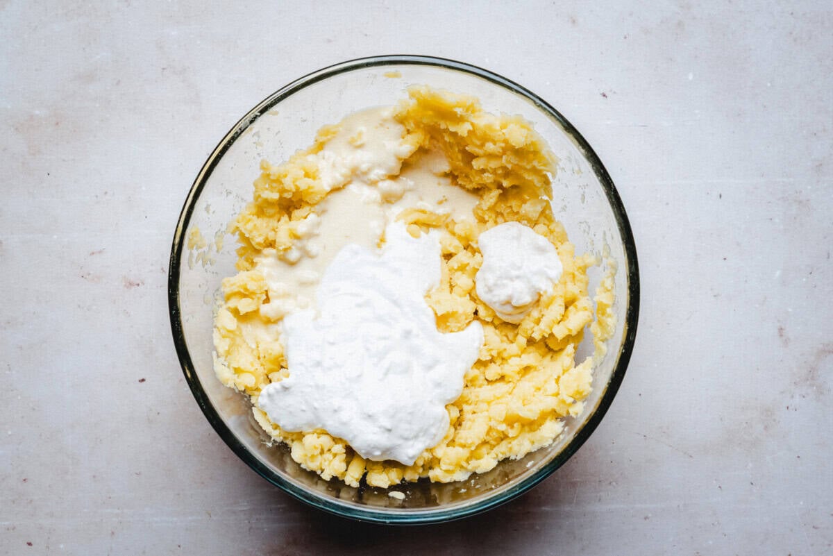 A glass bowl filled with mashed potatoes topped with a dollop of sour cream, sitting on a light-colored surface.