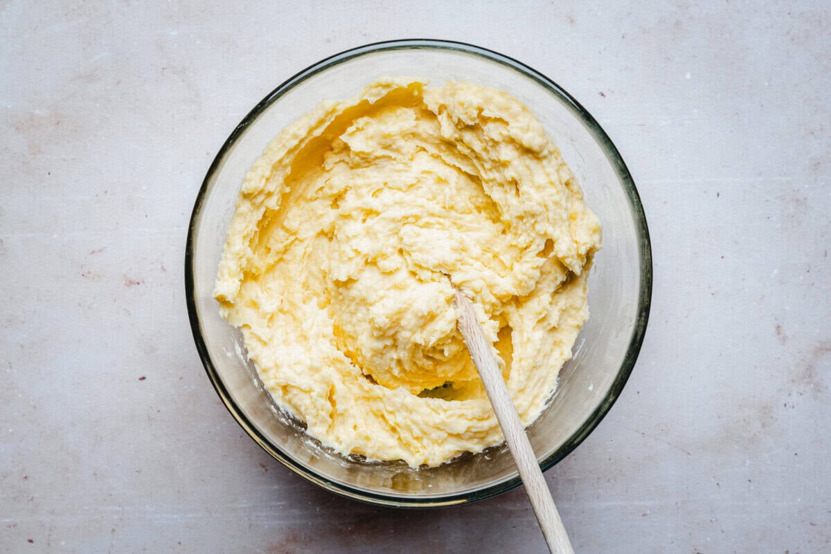 A glass bowl filled with a creamy, yellow mashed potatoes being mixed with a wooden spoon, placed on a light-colored countertop.