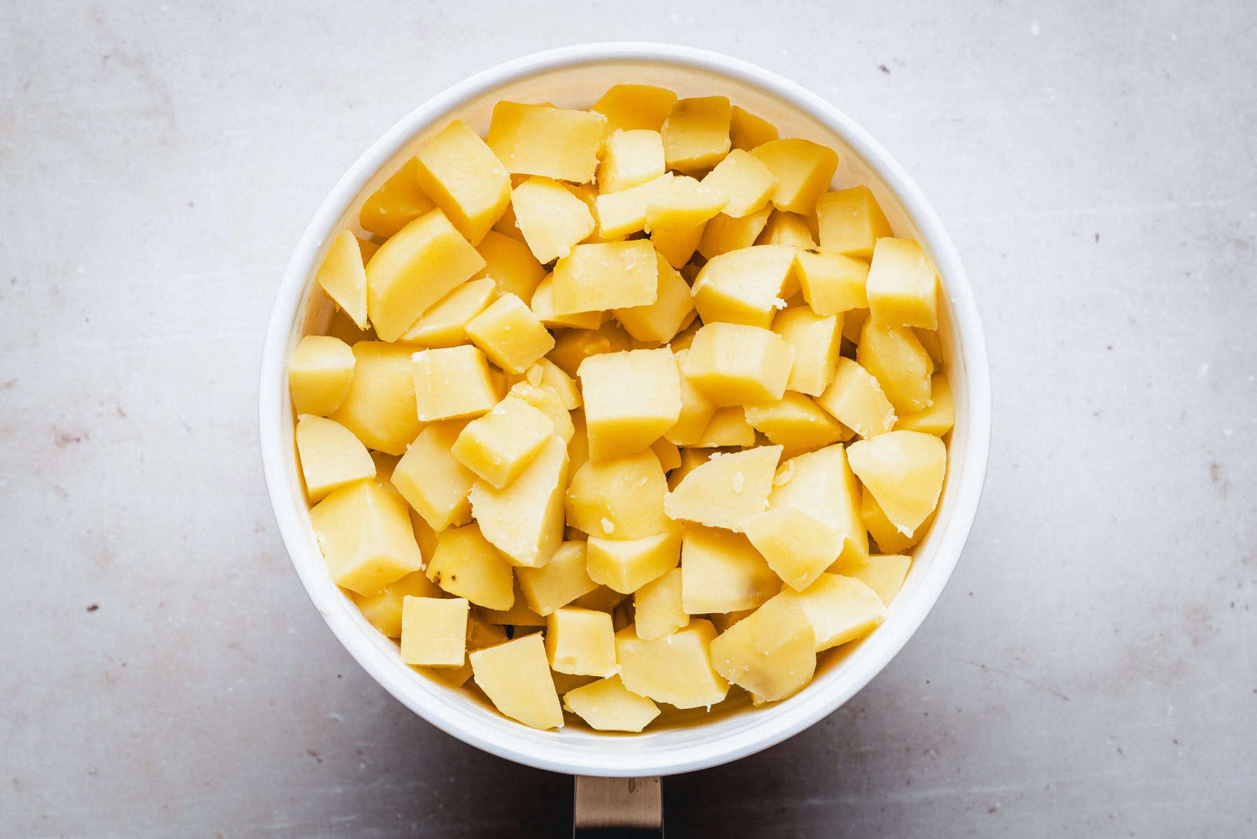 A bowl filled with evenly cut, peeled, and cooked potato cubes, viewed from above on a light-colored surface.