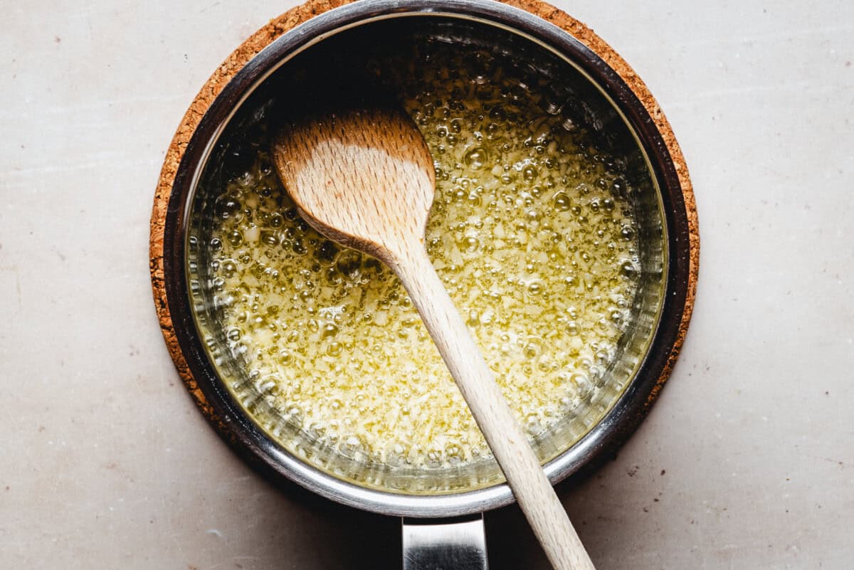 A wooden spoon rests in a saucepan filled with bubbling melted butter on a light-colored surface.