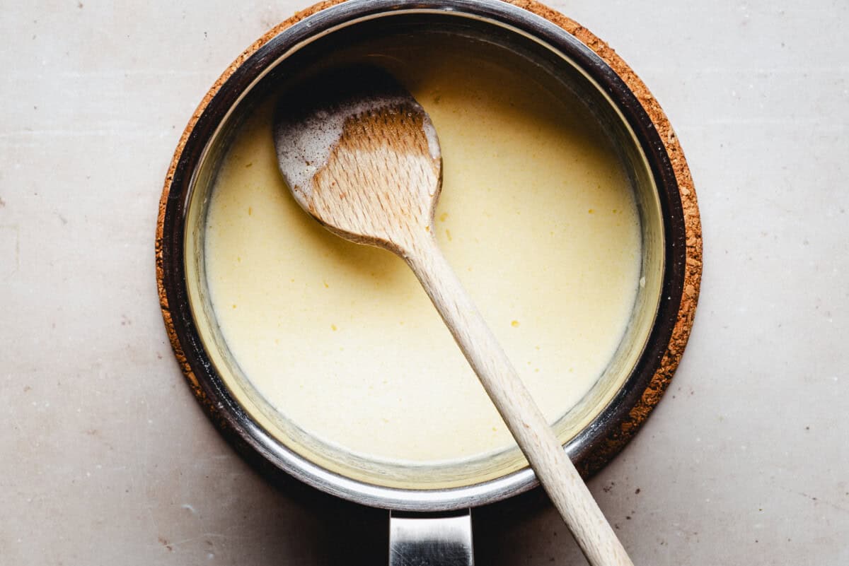 A saucepan filled with a creamy yellow sauce, with a wooden spoon resting inside, seen from above on a light-colored surface.
