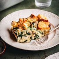 A slice of savory bread pudding with spinach, cheese, and herbs sits on a decorative white plate next to a fork. The bread cubes are golden and toasted, and the dish is served on a rustic green table.