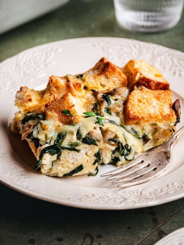 A slice of savory bread pudding with spinach, cheese, and herbs sits on a decorative white plate next to a fork. The bread cubes are golden and toasted, and the dish is served on a rustic green table.