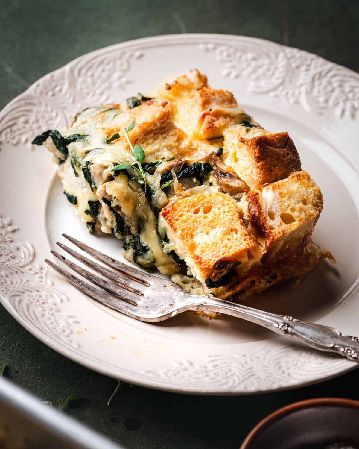 A slice of savory bread pudding with spinach, cheese, and mushrooms sits on a white decorative plate, accompanied by a fork. The dish features golden, toasted bread cubes and leafy greens.