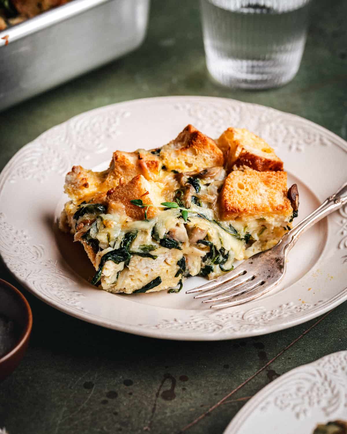 A slice of savory bread pudding with spinach, cheese, and mushrooms sits on a white plate with a vintage fork. A glass of water and more casserole are in the background.