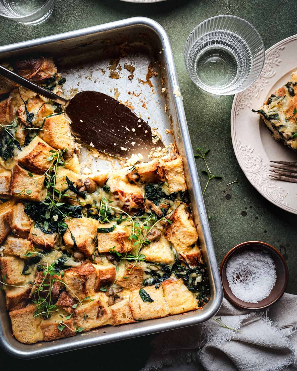 A rectangular baking dish with a savory bread pudding, topped with greens and herbs. A serving spatula rests in the dish, some pieces are missing. Nearby are a plate, fork, glasses, and a small bowl of salt.
