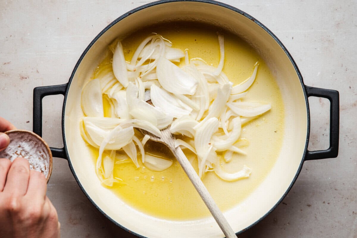 Sliced onions in a large pot with melted butter, being stirred with a wooden spoon. A hand holds a small bowl of salt over the pot, ready to season the onions.