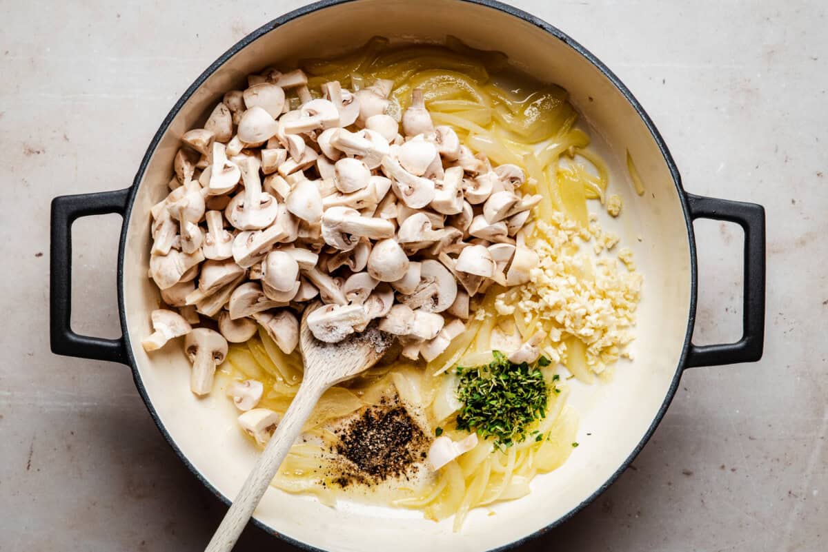 A large pot contains sliced onions, chopped mushrooms, minced garlic, chopped herbs, black pepper, and oil or butter, with a wooden spoon resting inside. The ingredients are ready to be cooked.