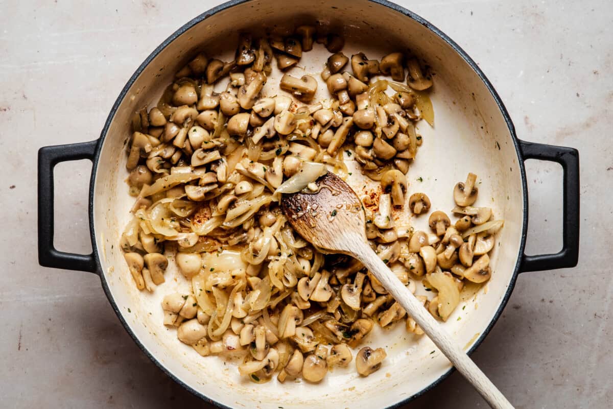 A large pot filled with sautéed mushrooms and onions, being stirred with a wooden spoon on a light countertop.