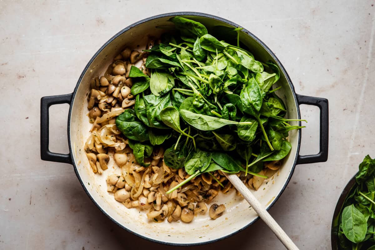 A large pot with sautéed mushrooms and onions on one side, and fresh spinach leaves piled on the other, with a wooden spoon resting inside. The pot sits on a light-colored countertop.