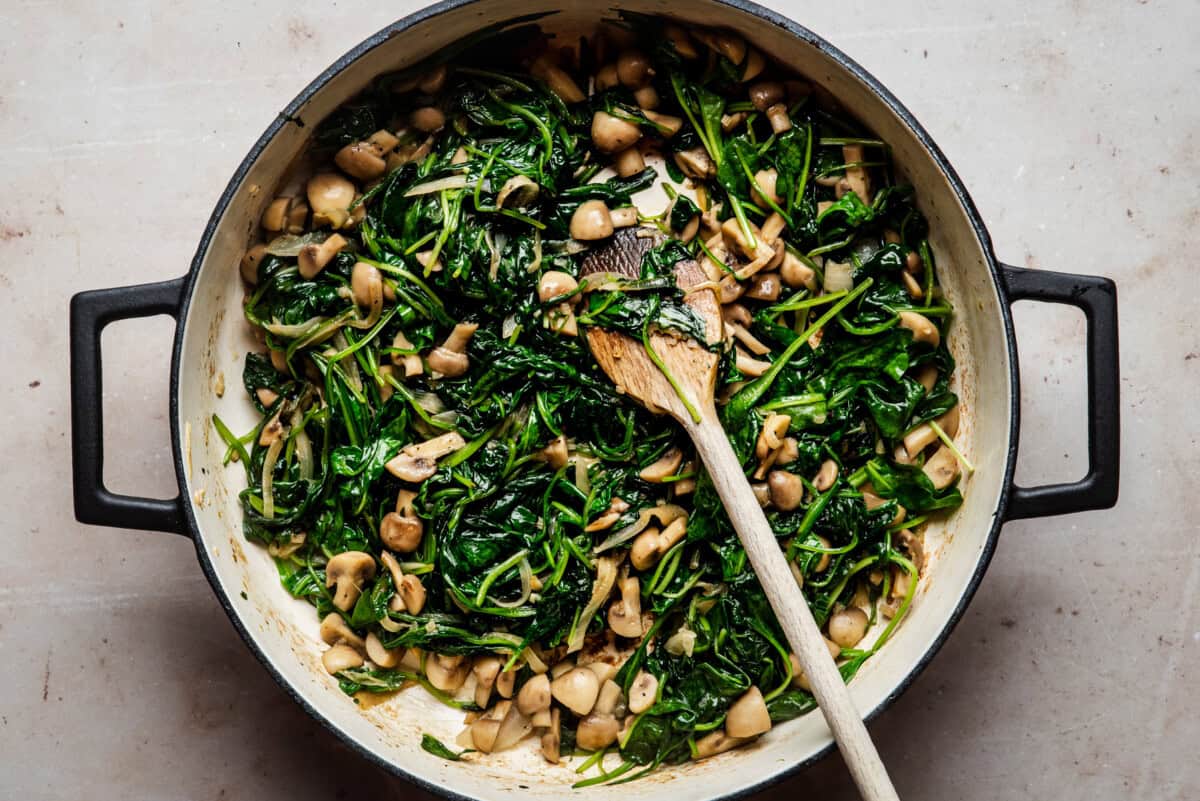A large pan filled with sautéed spinach and chopped mushrooms, mixed together and stirred with a wooden spoon on a light-colored surface.